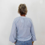 A person viewed from behind wearing a light blue long-sleeve blouse with a gathered neckline and blue jeans, standing against a plain white wall.