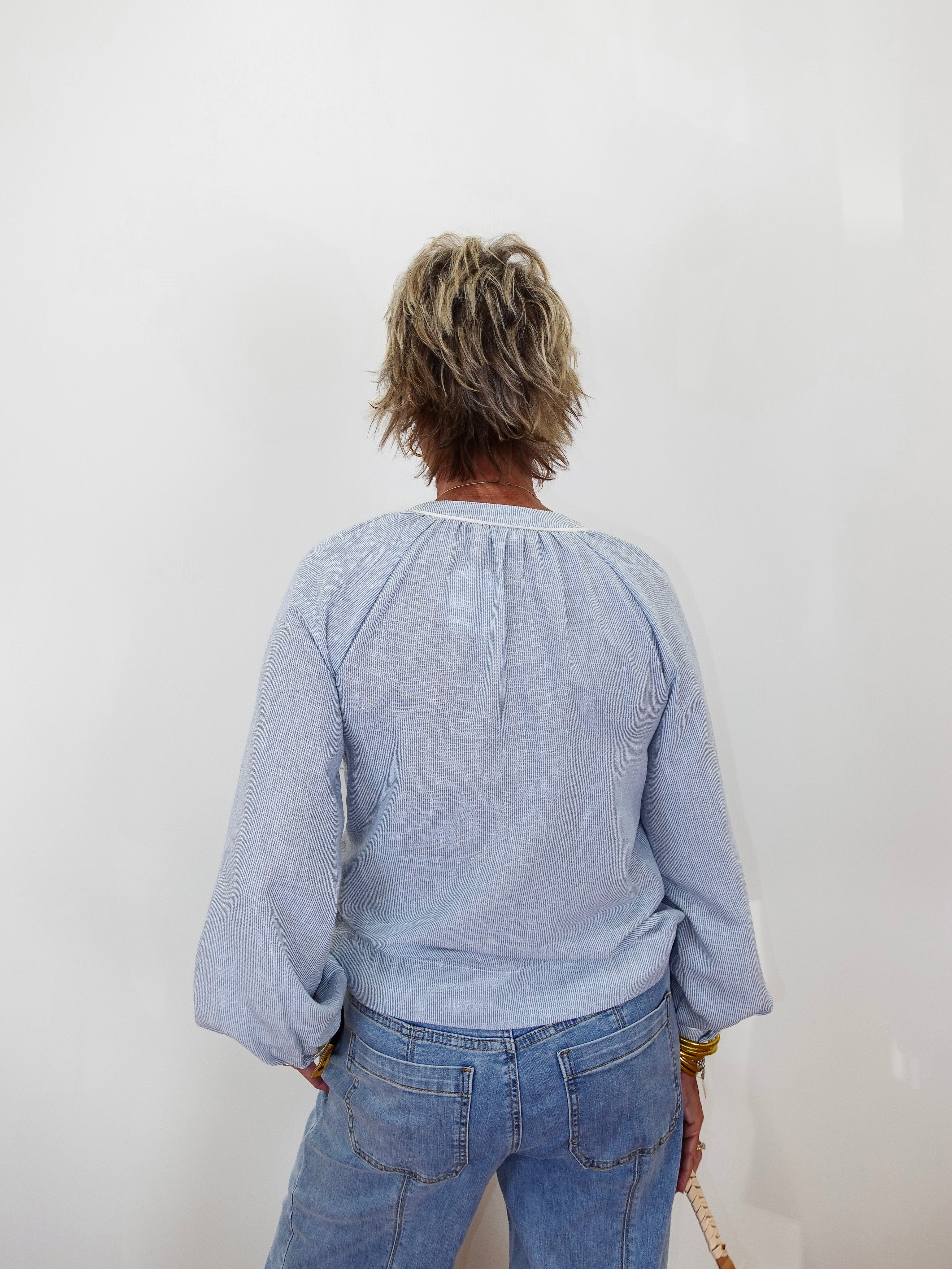 A person viewed from behind wearing a light blue long-sleeve blouse with a gathered neckline and blue jeans, standing against a plain white wall.