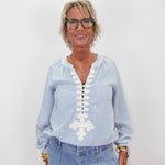 A woman with short blonde hair and glasses wearing a light blue long-sleeve blouse with white embroidered details down the front, paired with light blue jeans, standing against a plain white background.