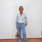 An older woman with short gray hair and glasses stands against a plain white wall wearing a light blue long-sleeve button-up shirt with white embroidery down the front, light-wash blue jeans, and beige sneakers. She is holding a tan handbag.