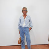 An older woman with short gray hair and glasses stands against a plain white wall wearing a light blue long-sleeve button-up shirt with white embroidery down the front, light-wash blue jeans, and beige sneakers. She is holding a tan handbag.