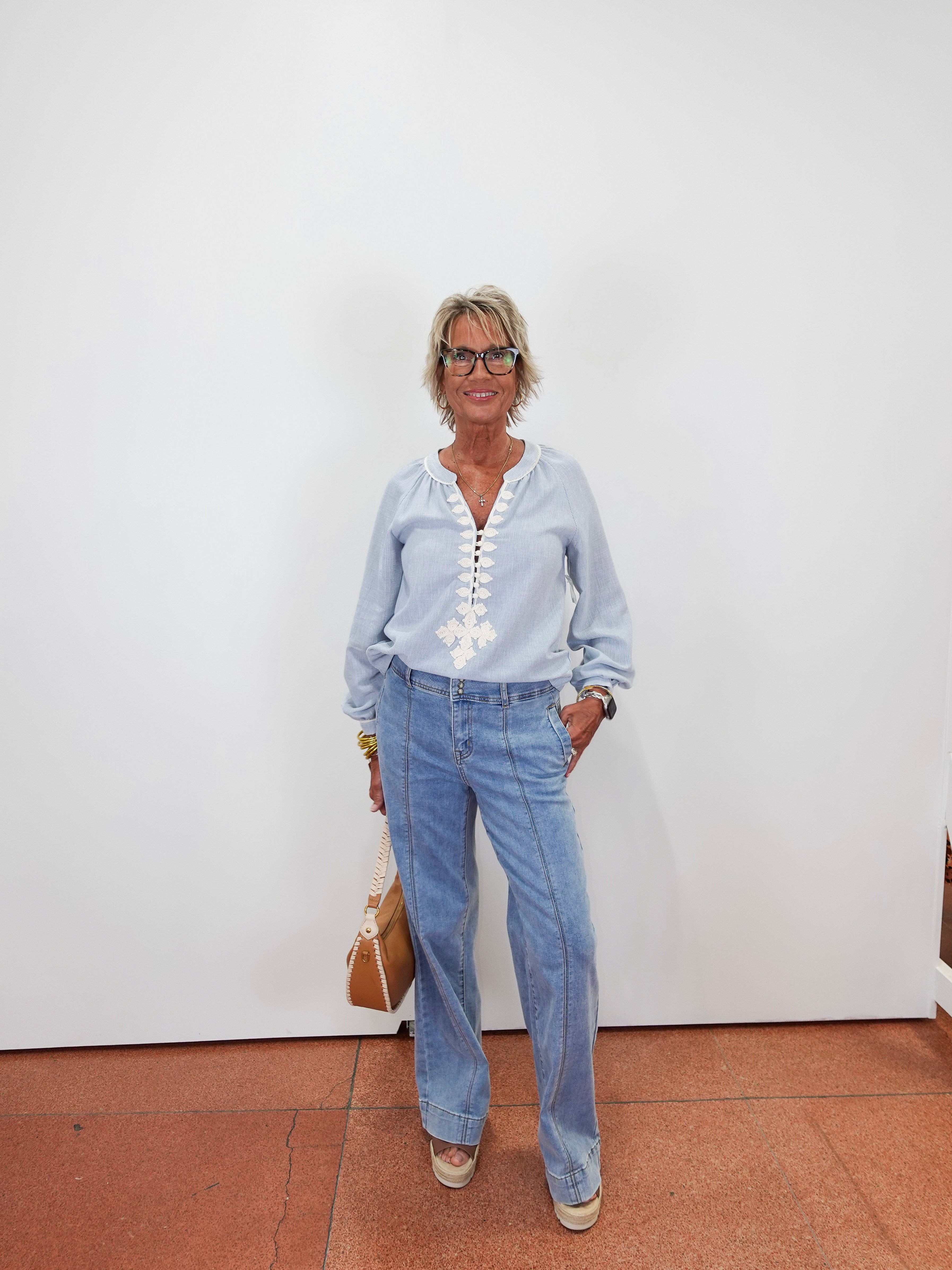 An older woman with short gray hair and glasses stands against a plain white wall wearing a light blue long-sleeve button-up shirt with white embroidery down the front, light-wash blue jeans, and beige sneakers. She is holding a tan handbag.
