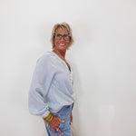 A smiling older woman with short gray hair and glasses stands against a plain white wall, wearing a light blue long-sleeve top with white lace trim and light blue jeans.