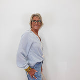 A smiling older woman with short gray hair and glasses stands against a plain white wall, wearing a light blue long-sleeve top with white lace trim and light blue jeans.