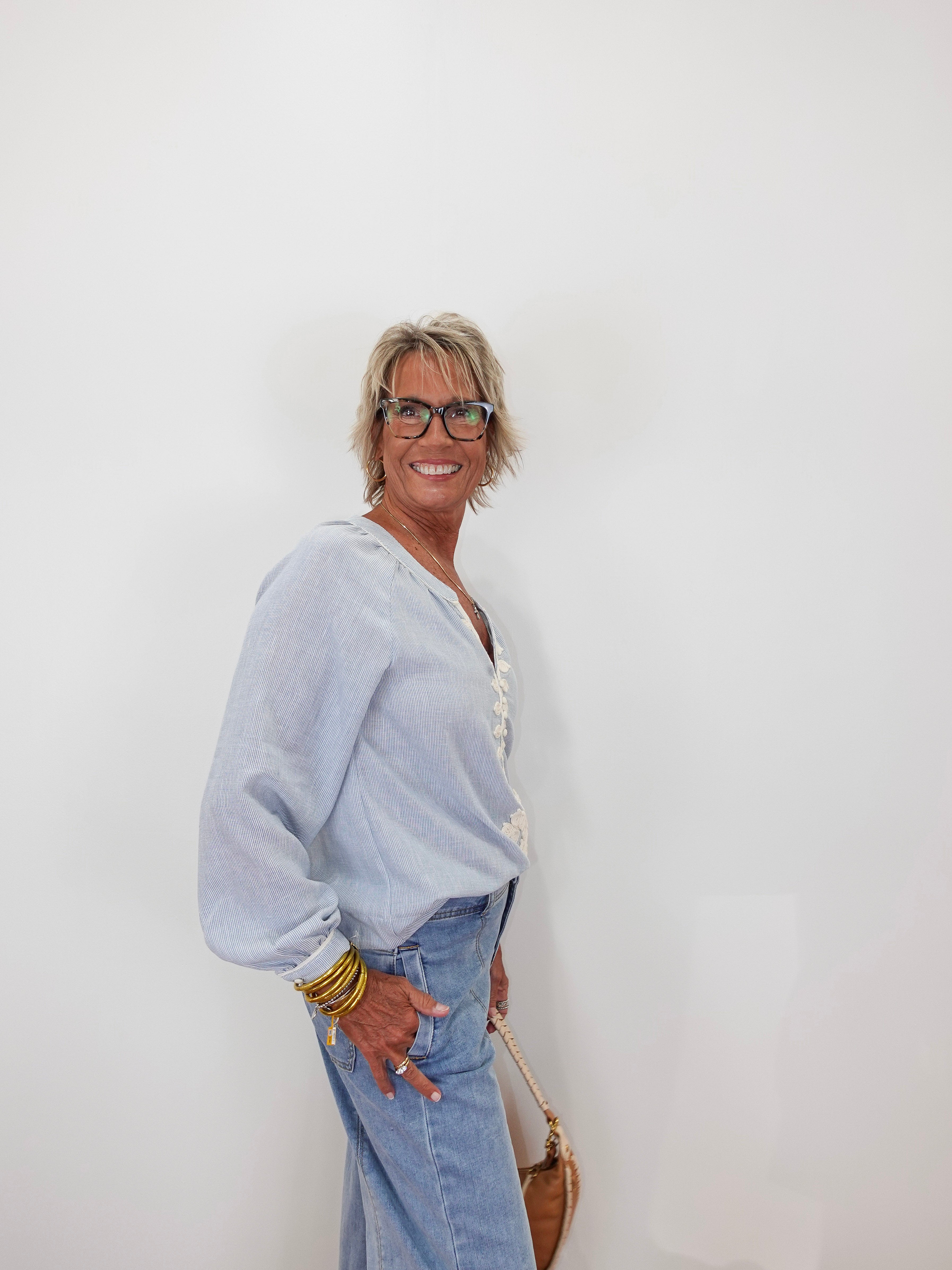 A smiling older woman with short gray hair and glasses stands against a plain white wall, wearing a light blue long-sleeve top with white lace trim and light blue jeans.