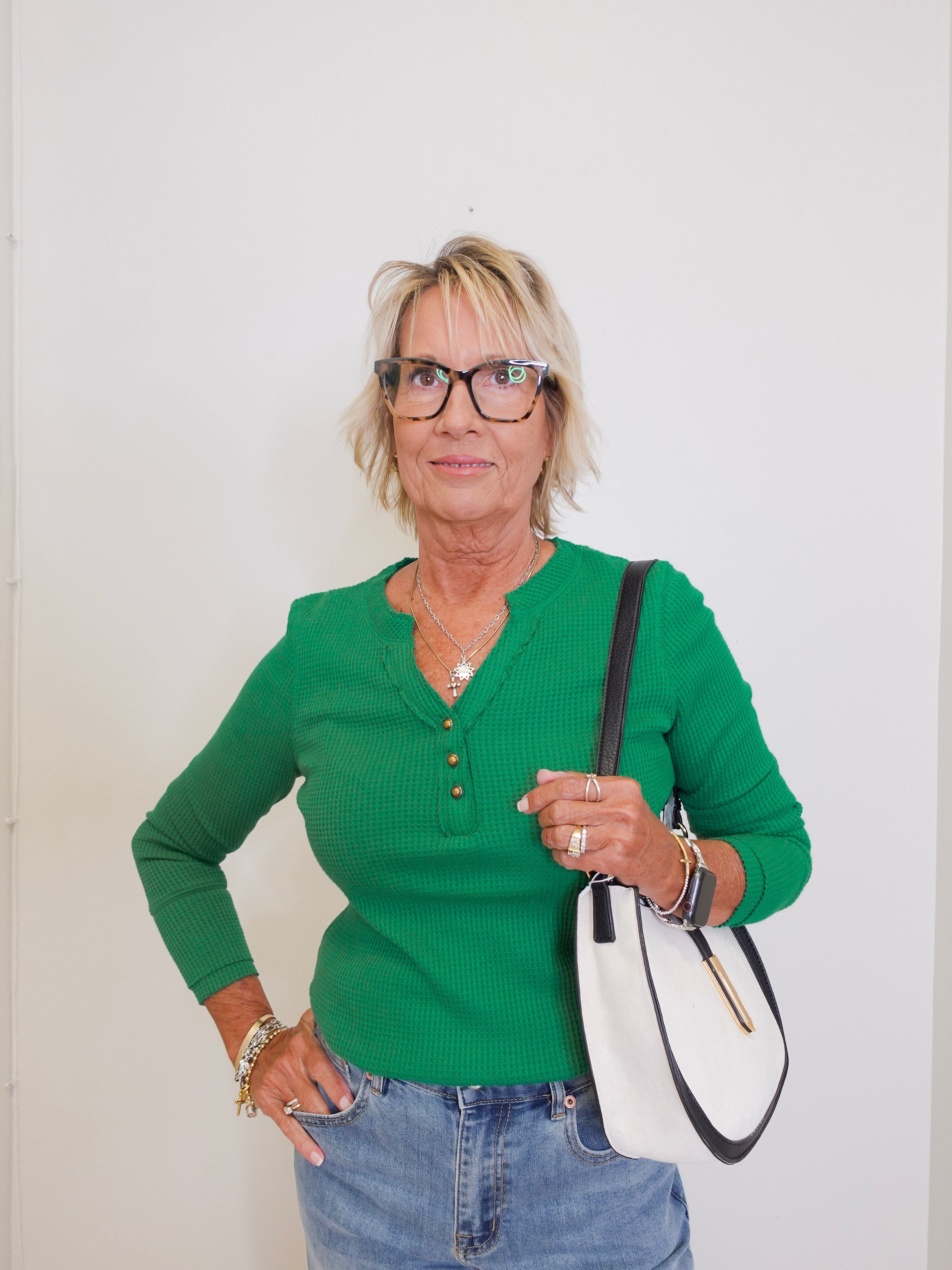 Woman wearing a green sweater and blue jeans, holding a white and black handbag against a white background.