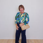 Woman wearing a colorful blouse and dark pants, holding a beige handbag against a white wall.