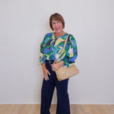 Woman wearing a colorful blouse and dark pants, holding a beige handbag against a white wall.