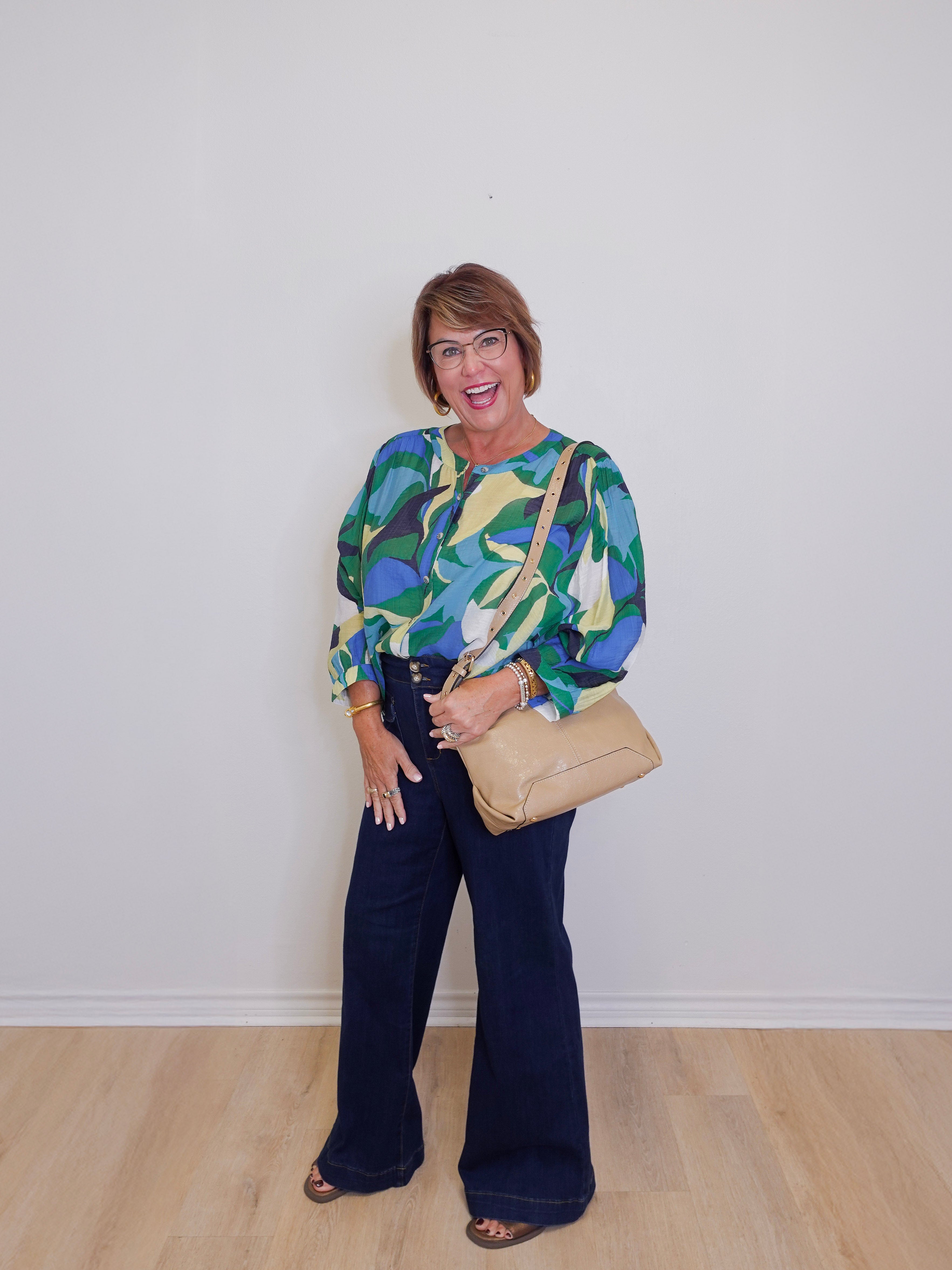 Woman wearing a colorful blouse and dark pants, holding a beige handbag against a white wall.