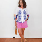 Woman wearing a white blouse with blue floral patterns and pink shorts against a white wall.