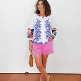 Woman wearing a white blouse with blue floral patterns and pink shorts against a white wall.