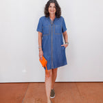Woman wearing a blue denim dress holding an orange clutch against a white wall.