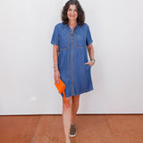 Woman wearing a blue denim dress holding an orange clutch against a white wall.