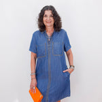 Woman wearing a blue denim dress holding an orange clutch against a white background