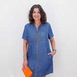 Woman wearing a blue denim dress holding an orange clutch against a white background