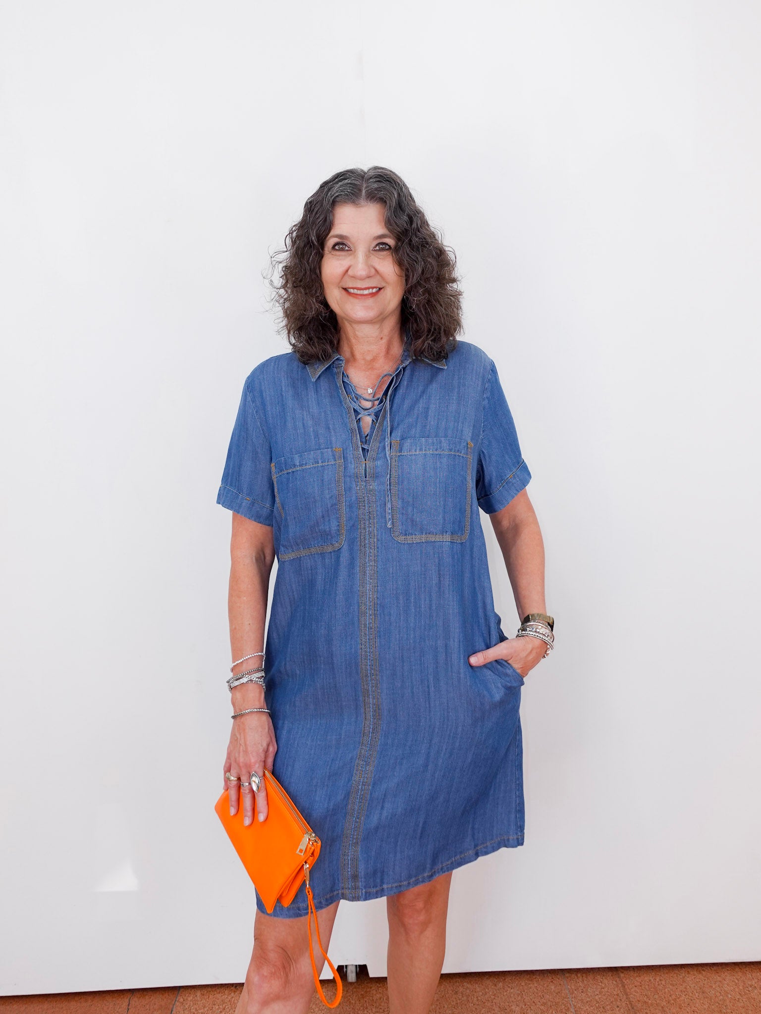 Woman wearing a blue denim dress holding an orange clutch against a white background