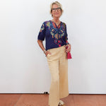 Woman wearing a colorful blouse and beige pants against a white wall.