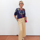Woman wearing a colorful blouse and beige pants against a white wall.