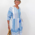 Woman wearing a blue and white striped dress holding a beige bag against a white wall.