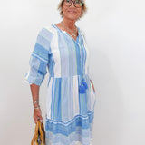 Woman wearing a blue and white striped dress holding a beige bag against a white wall.