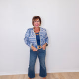 Woman wearing a blue patterned jacket, blue jeans, and brown shoes against a white wall.