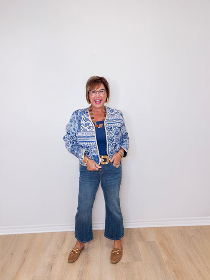 Woman wearing a blue patterned jacket, blue jeans, and brown shoes against a white wall.