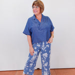 Woman wearing a blue shirt and floral pants standing against a white wall.