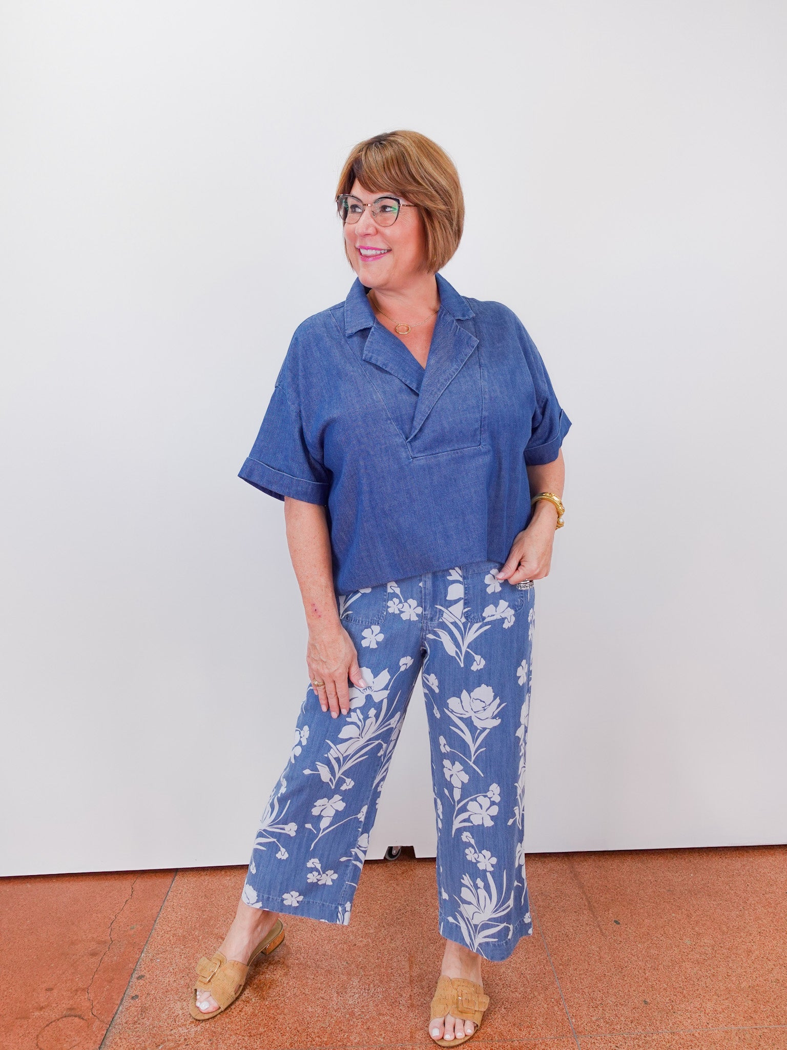 Woman wearing a blue shirt and floral pants standing against a white wall.