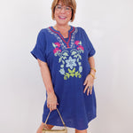 Woman wearing a blue embroidered dress holding a gold handbag against a white background