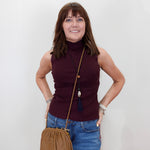 A woman with brown hair and bangs stands against a white wall wearing a sleeveless ribbed turtleneck top in deep burgundy, blue jeans, and a tan leather crossbody bag.