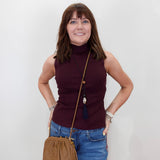 A woman with brown hair and bangs stands against a white wall wearing a sleeveless ribbed turtleneck top in deep burgundy, blue jeans, and a tan leather crossbody bag.