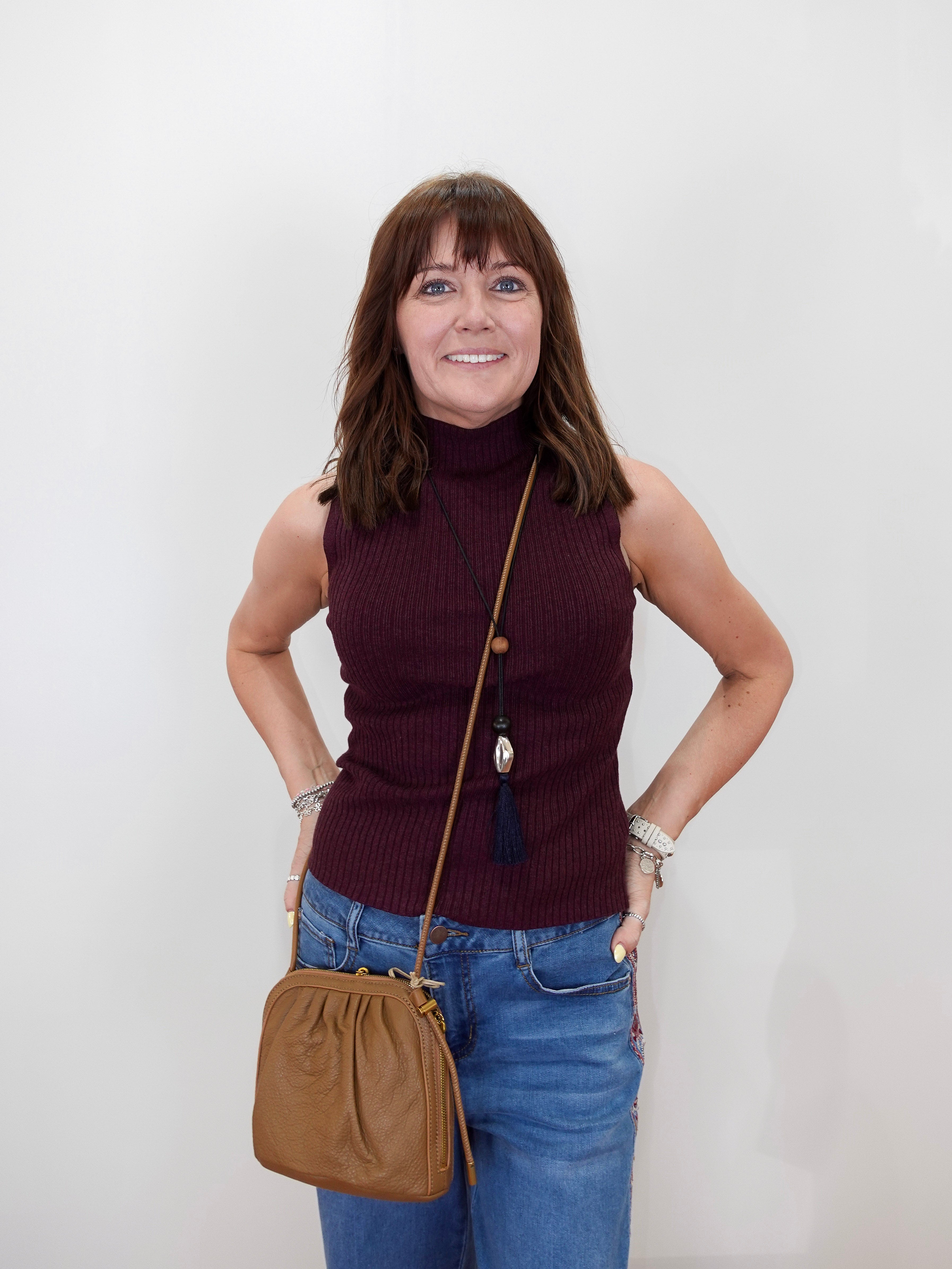 A woman with brown hair and bangs stands against a white wall wearing a sleeveless ribbed turtleneck top in deep burgundy, blue jeans, and a tan leather crossbody bag.