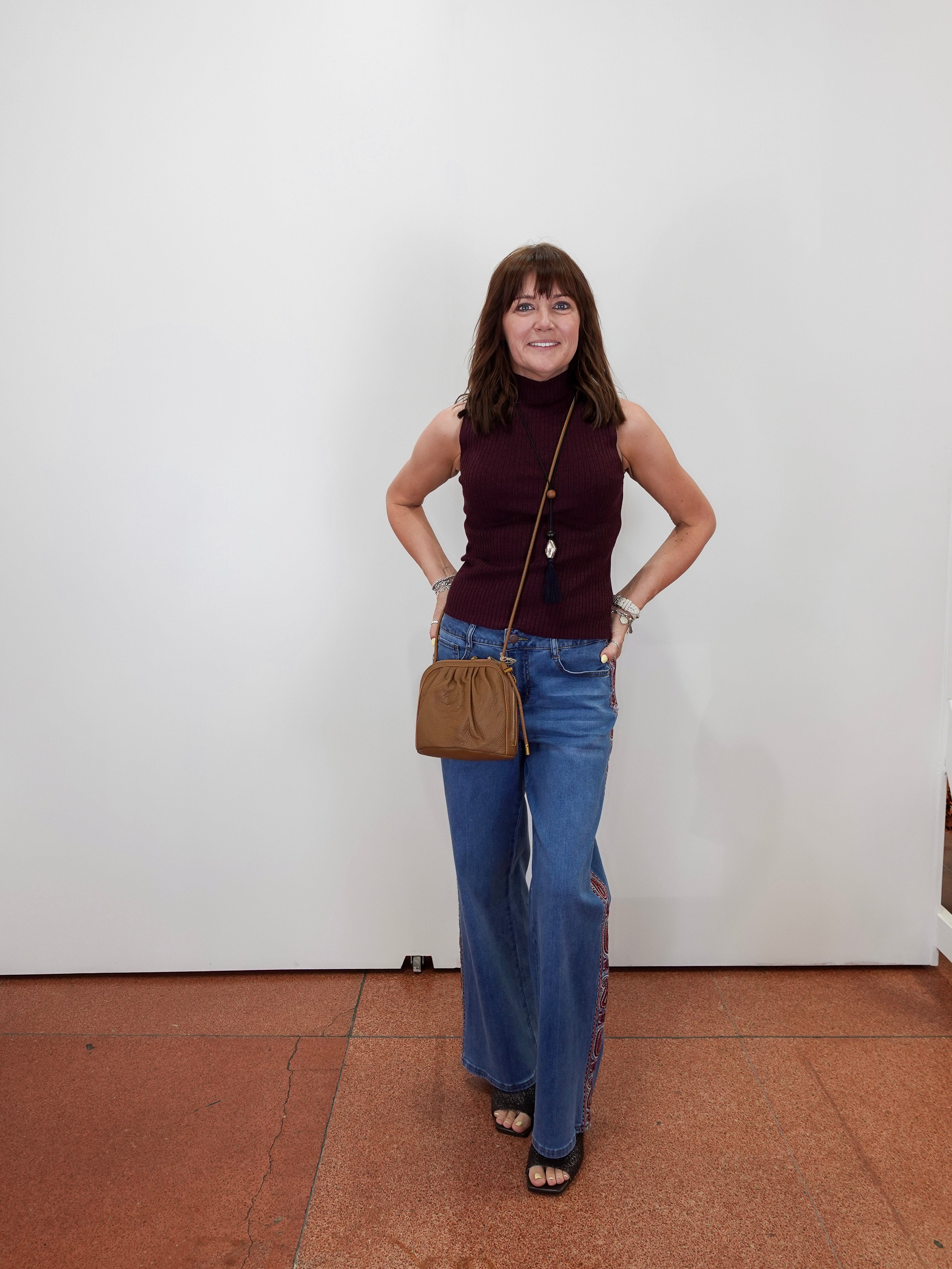 A woman stands against a plain white wall wearing a sleeveless burgundy mock-neck top, blue wide-leg jeans, and black sandals, while carrying a brown crossbody bag.