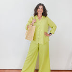 Woman in a bright green outfit holding a woven bag against a white wall.