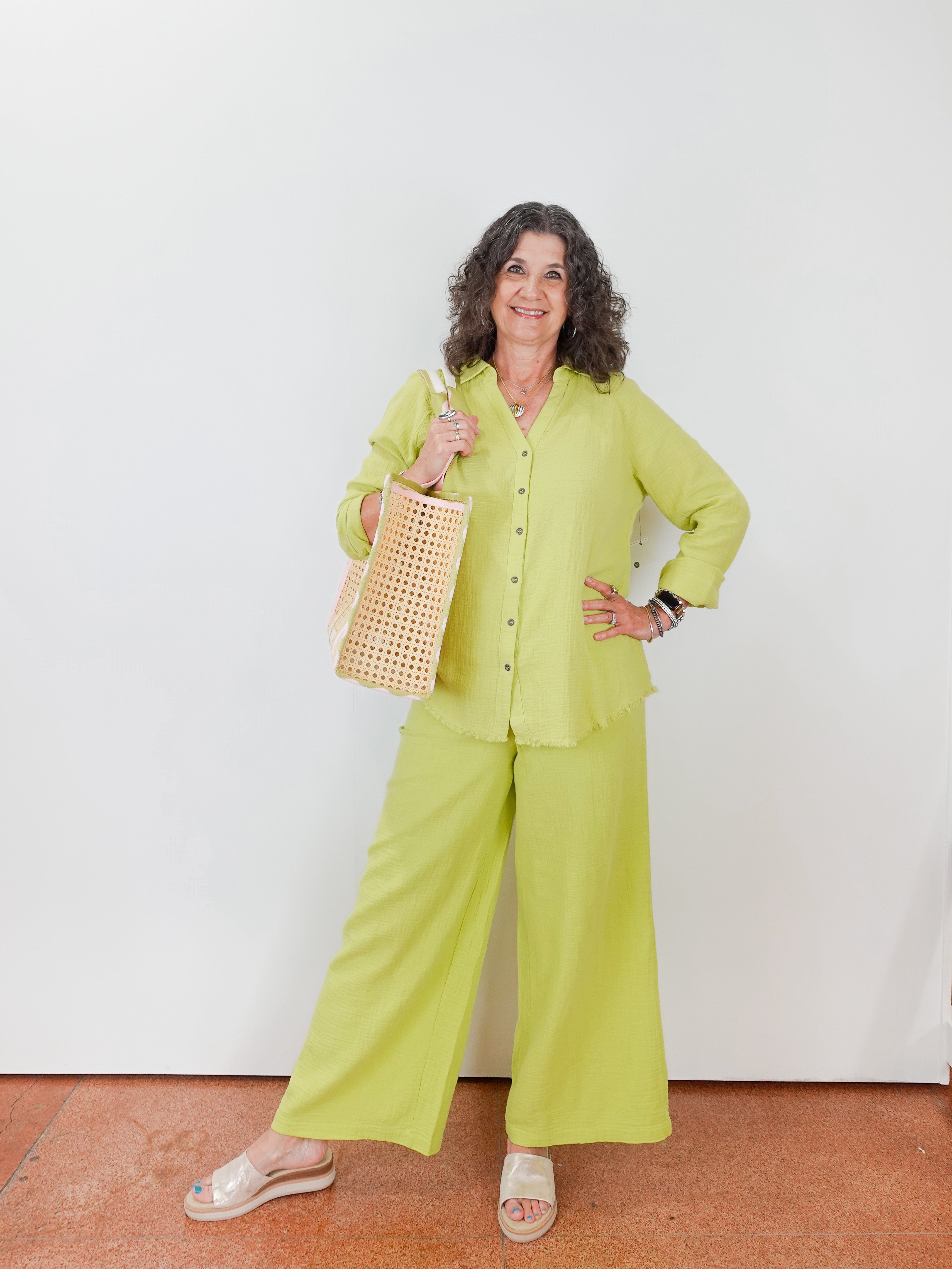 Woman in a bright green outfit holding a woven bag against a white wall.
