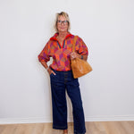 Person wearing a colorful patterned shirt and blue jeans, holding a brown leather bag against a white wall.