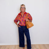 Person wearing a colorful patterned shirt and blue jeans, holding a brown leather bag against a white wall.