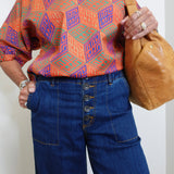 Person wearing a colorful geometric patterned shirt and blue jeans, holding a brown bag.