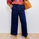 Person wearing a colorful kimono-style top, blue wide-leg jeans, and tan shoes, holding a brown leather bag against a white wall.