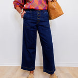 Person wearing a colorful kimono-style top, blue wide-leg jeans, and tan shoes, holding a brown leather bag against a white wall.