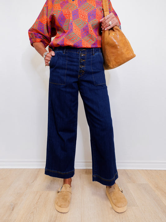 Person wearing a colorful kimono-style top, blue wide-leg jeans, and tan shoes, holding a brown leather bag against a white wall.