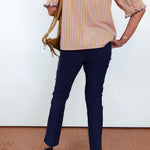 A person shown from the back wearing a beige and orange vertically striped blouse with ruffled sleeves, navy blue pants, and black wedge sandals, while carrying a tan shoulder bag against a white wall.