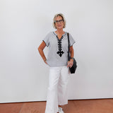 Woman wearing a gray top with black lace details, white pants, and white sneakers against a white wall.