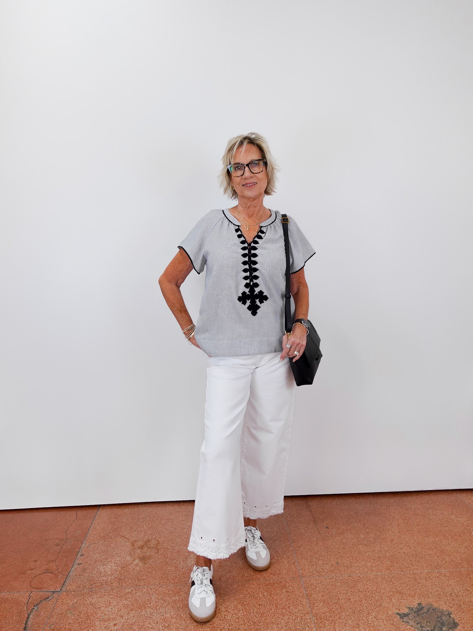 Woman wearing a gray top with black lace details, white pants, and white sneakers against a white wall.