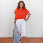 Woman wearing an orange shirt and white pants, holding a blue jacket against a white wall.