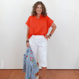 Woman wearing an orange shirt and white pants, holding a blue jacket against a white wall.