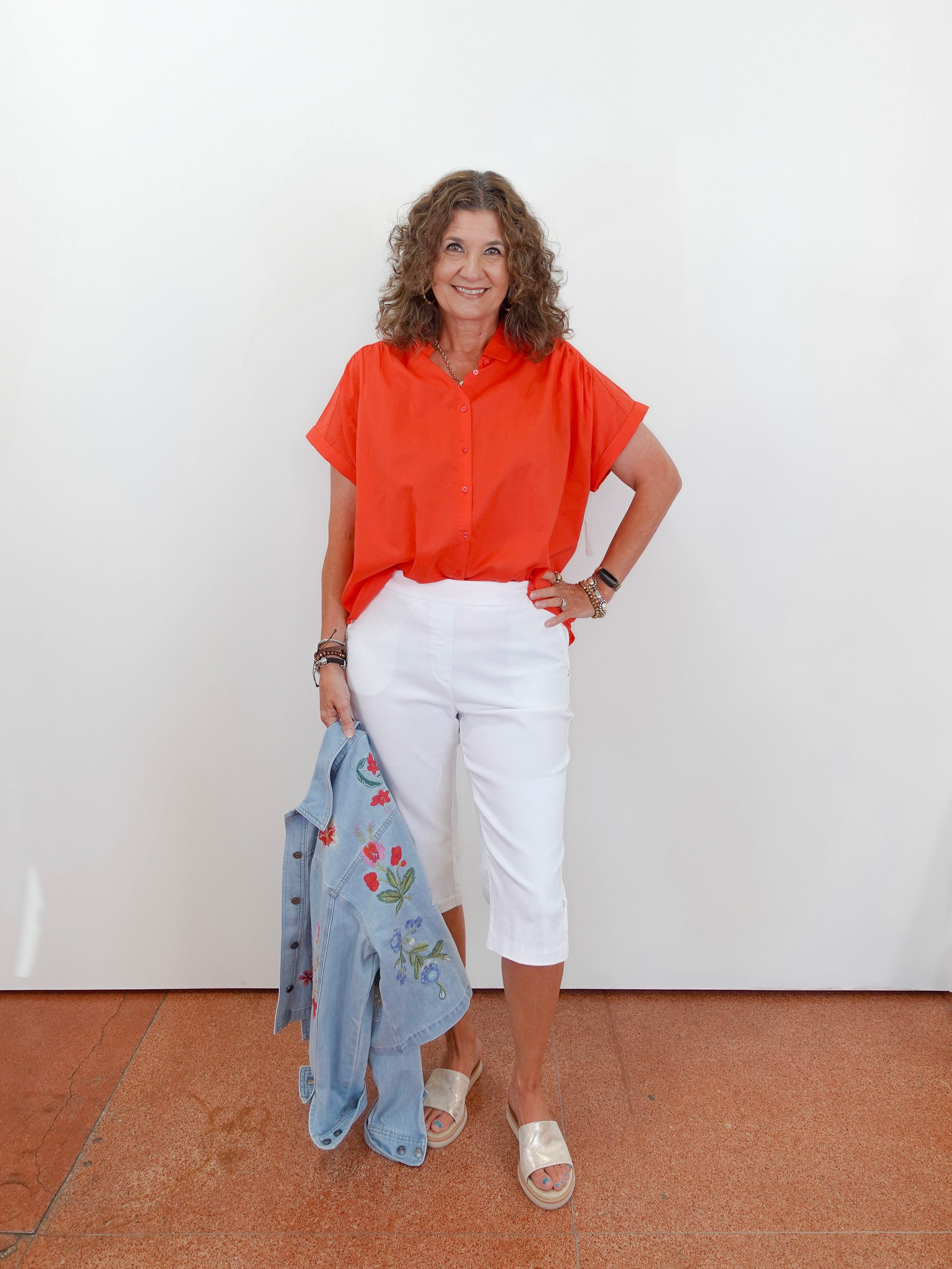 Woman wearing an orange shirt and white pants, holding a blue jacket against a white wall.