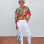 Woman wearing a colorful patterned shirt and white pants, standing against a plain background.