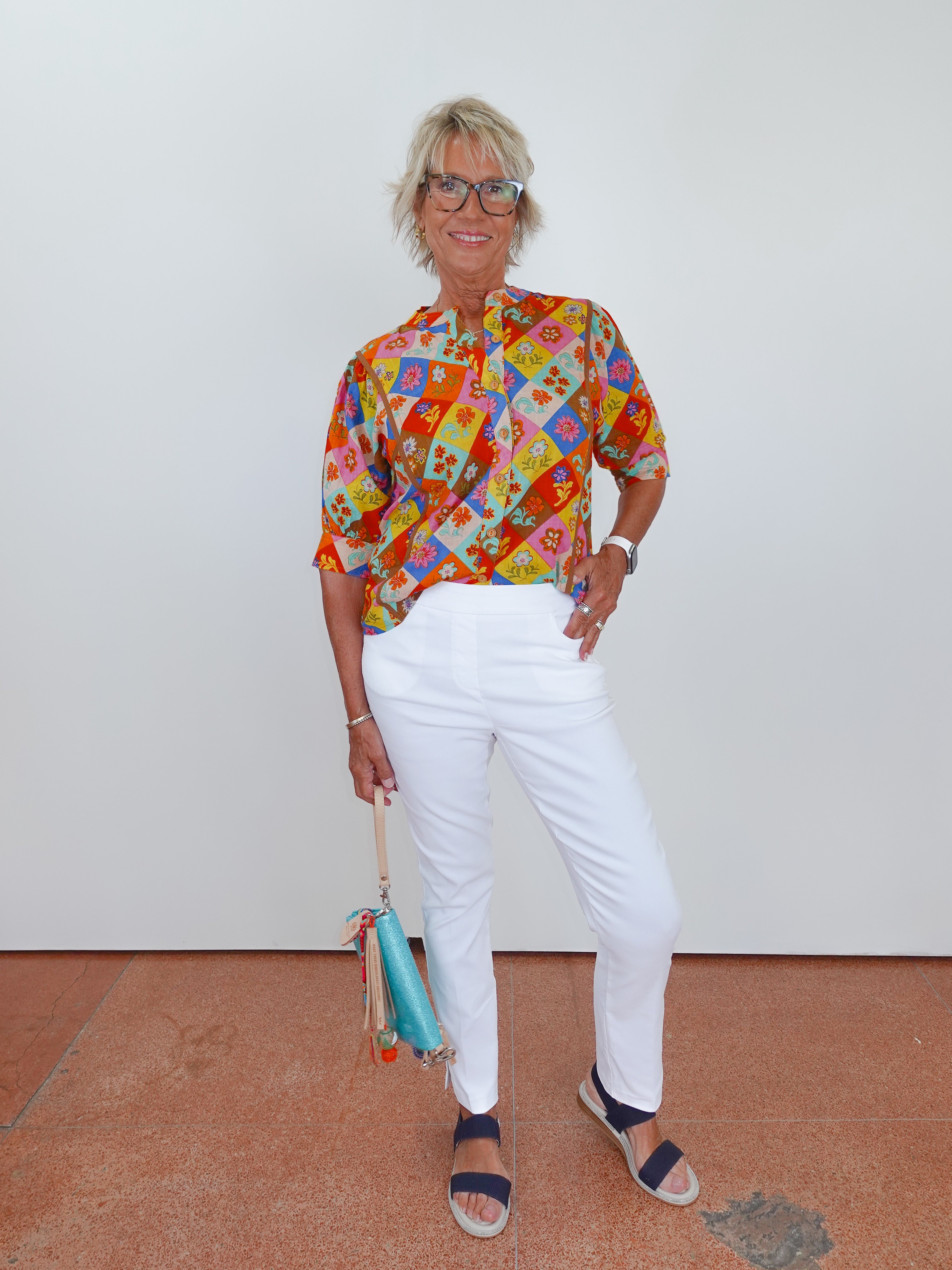 Woman wearing a colorful patterned shirt and white pants, standing against a plain background.