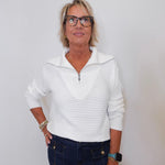 Woman wearing a white sweater and dark pants against a plain background